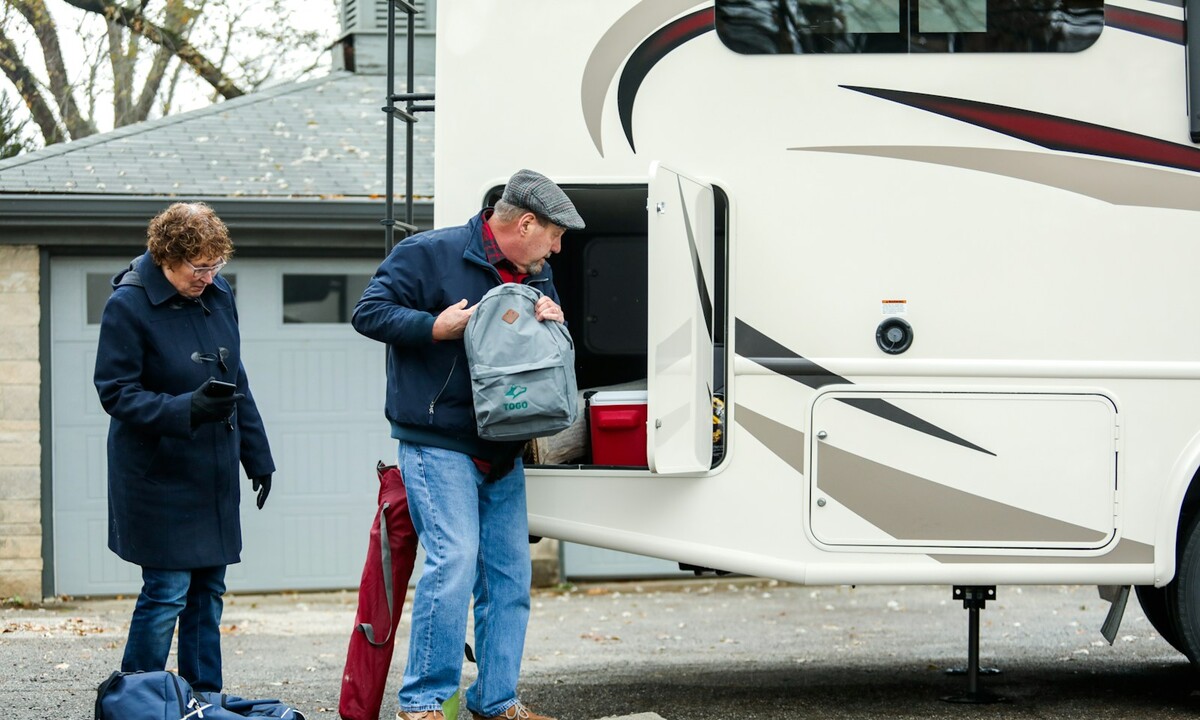 A couple loading a backpack, cooler and bags into the outdoor storage bay of a motorhome before a trip