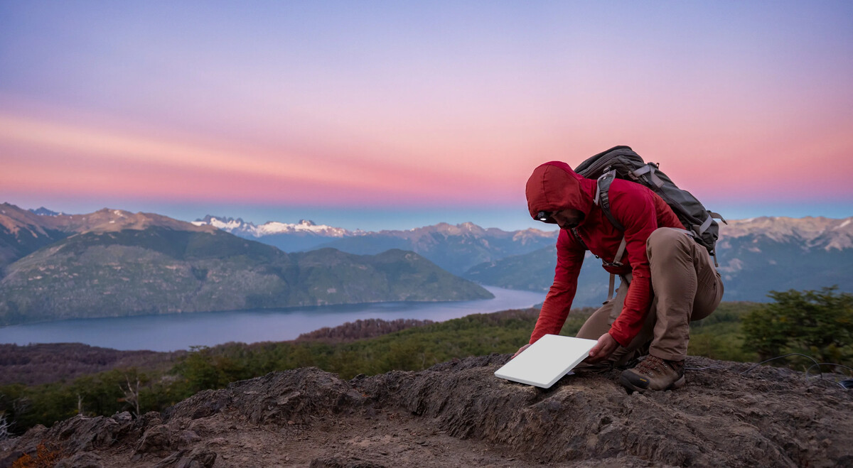 A hiker setting up a Starlink Mini dish on a mountain ridge at sunset, with a lake and snow-capped peaks in the background